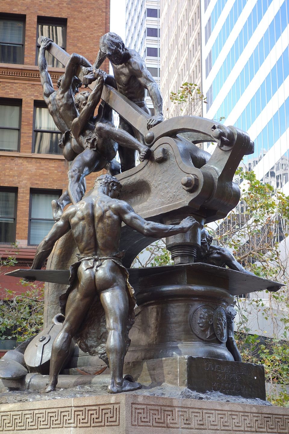 Douglas Tilden's bronze Mechanics Monument: muscular workers straining at a giant punch press, on a granite base carved with a Greek key, with downtown towers behind.